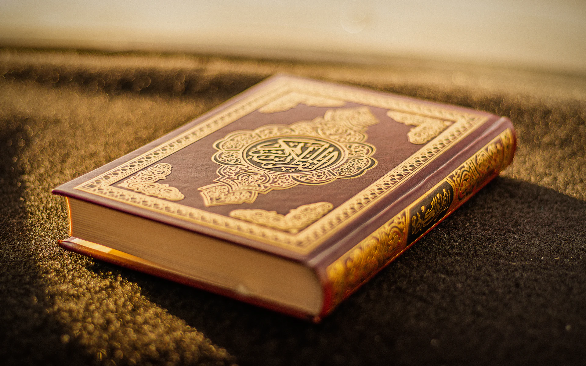 A Quran with a gold-patterned cover resting on a prayer mat in warm light
